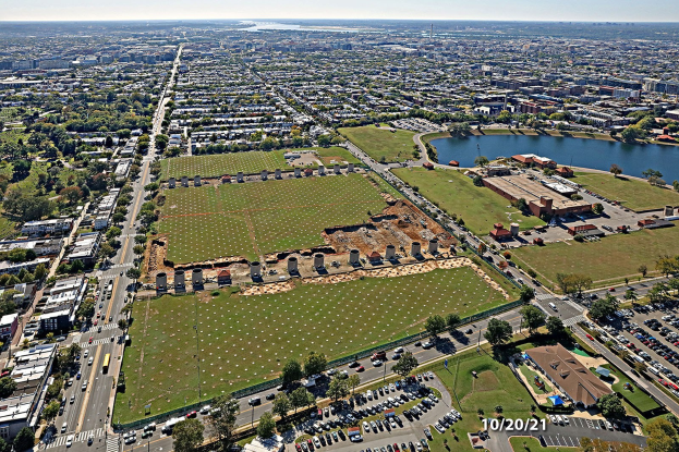 Luftaufnahme einer Stadt mit Gebäuden, Bäumen, Straßen, Fahrzeugen, Wasser und Wolken, die einen großen zentralen Platz zeigt, der als Park geplant ist.