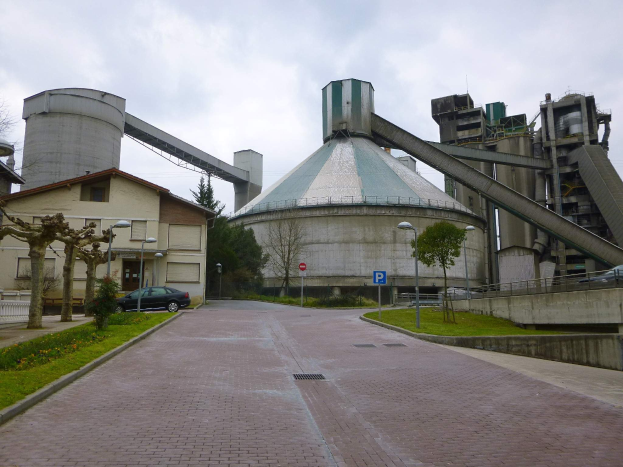 Große Zementfabrik mit Gebäuden, Fahrzeugen und Straßeninfrastruktur entlang einer Straße unter einem bewölkten Himmel.