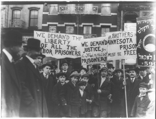 Schwarze und weiße Fotografie einer Gruppe von Menschen in einer Reihe mit Schildern, die "Wir fordern die Freiheit aller Gerechtigkeit für die Gefangenen" tragen, vor einem Gebäude mit Fenstern und einem Geländer.