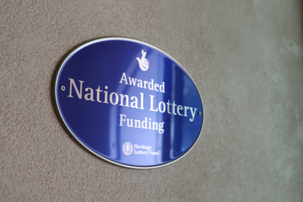 Blauer rechteckiger Schild an einem Gebäude mit weißem Hintergrund und schwarzer fetter Schrift, die "National Lottery Funding" liest.