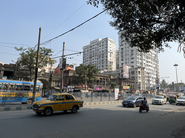 Gelbes Taxi auf einer belebten Straße in Mumbai mit hohen Gebäuden, Bäumen, Strommasten mit Drähten, Laternen und Texttafeln vor einem sichtbaren Himmel.
