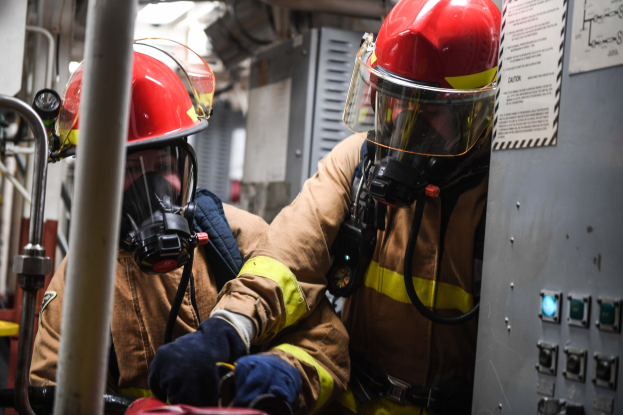 Zwei Feuerwehrleute in Schutzausrüstung arbeiten an einem Feuerwehrauto, mit einer Tafel mit Text rechts und Metallstangen im Hintergrund.