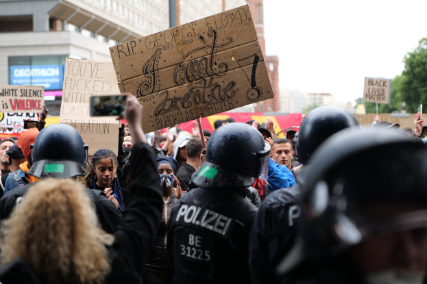 Eine Gruppe von Menschen in Helmen mit Protestschildern und Mobiltelefonen vor einer Menge, mit Gebäuden, Bäumen und einem Pfahl im Hintergrund.