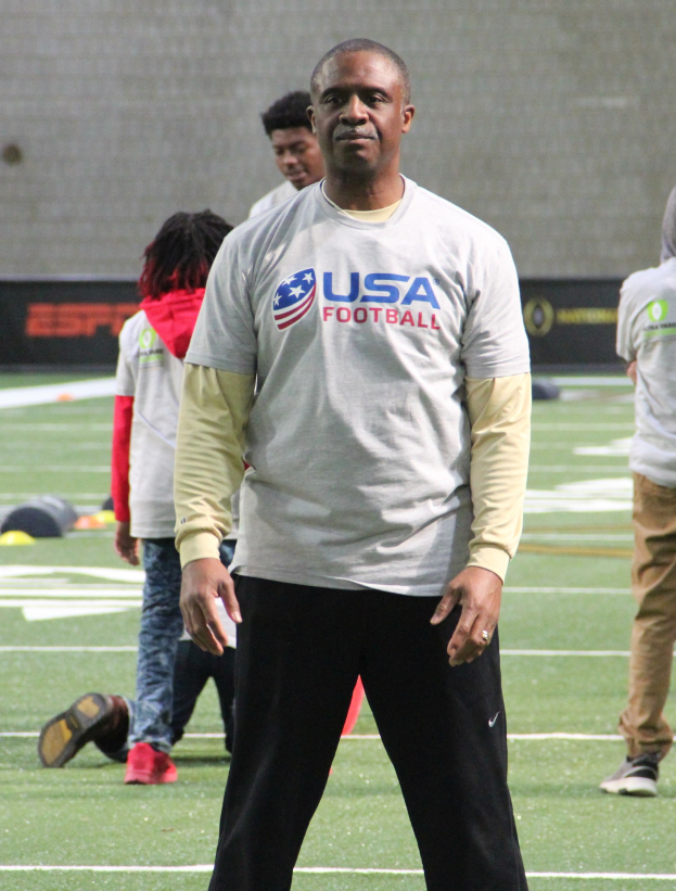 Ein Fußballspieler in Stollenschuhen steht auf einem Feld mit einer Gruppe von Menschen und einer Wand im Hintergrund.