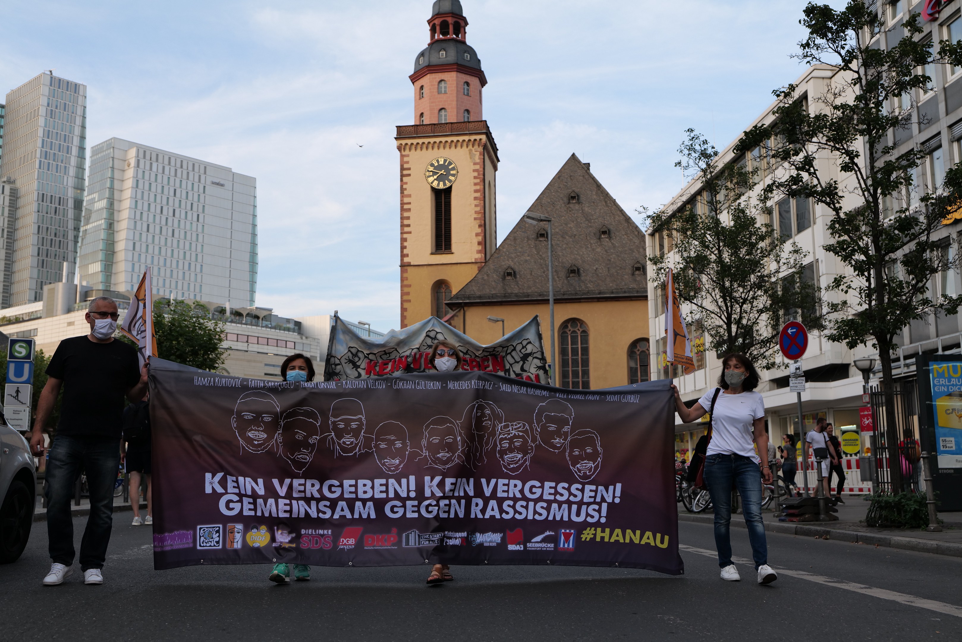 Gruppe von maskierten Menschen, die eine Plakattafel tragen, auf einer Straße mit einem geparkten Auto auf der linken Seite, Gebäuden, Bäumen und einer Kirchturmuhr im Hintergrund und einem klaren blauen Himmel.
