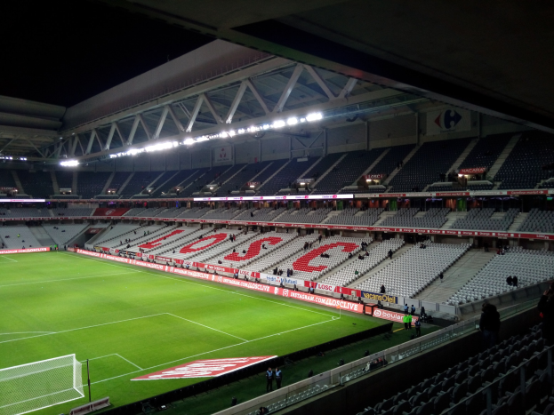 Ein großes Stadion mit einem gut beleuchteten Fußballfeld, das grüne Rasen, ein Netz auf der linken Seite und einige Zuschauer in den Rängen zeigt, vor einem dunklen Hintergrund.