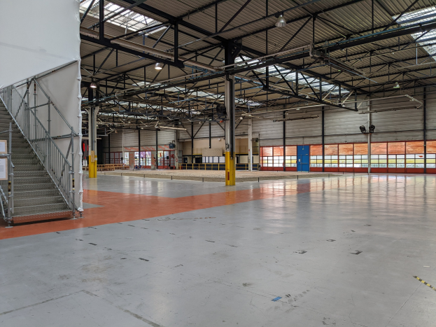Große Industriehalle mit zahlreichen Fenstern, Metallträgern, einer Treppe mit Geländer, einem Schild, Deckenleuchten und zusätzlichen Trägern.