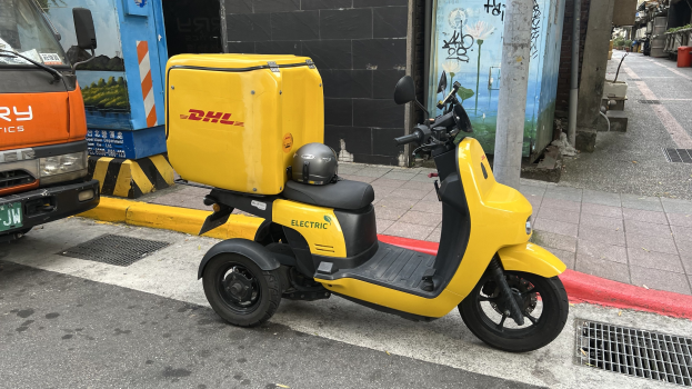 Ein gelbes Elektro-Scooter mit Helm darauf auf einer Straße neben einem Lastwagen geparkt, mit Menschen auf einem Fußweg und Gebäuden im Hintergrund.