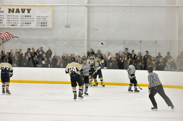 Gruppe von Menschen, die Eis-hockey auf einem Eis-hockeyfeld spielen, mit Zuschauern auf den Tribünen und einem Banner im Hintergrund.