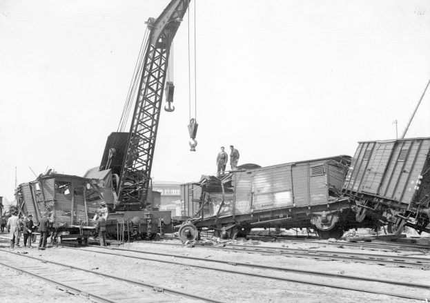 Schwarzes und weißes Bild einer Zugkatastrophe mit einem Kran, der einen Zugwagen auf die Schienen hebt, mehrere Menschen in der Nähe, Gebäude und einen klaren Himmel im Hintergrund.