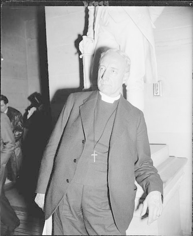 Schwarzes und weißes Foto von Papst Johannes Paul II. in Anzug und Krawatte, der neben einer Statue steht, mit ein paar Menschen und einer Wand im Hintergrund.