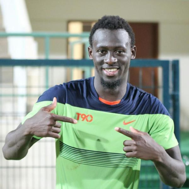 Ein Mann in einem grün-blauen Hemd lächelt, waehrend er auf die Kamera zeigt, mit einem unscharfen Geländer, Wand und Tür im Hintergrund.