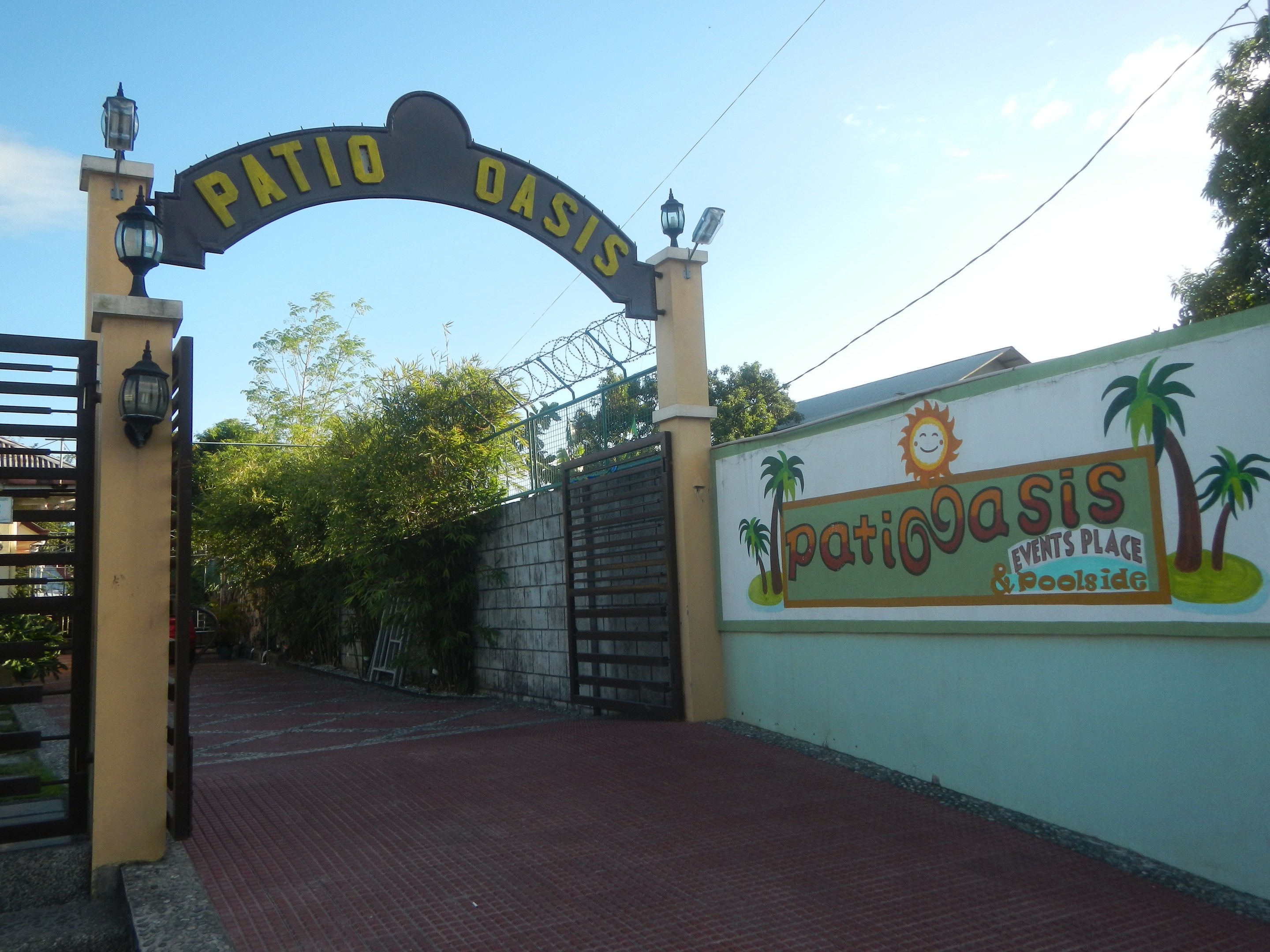 Eintritt zum Patio Oasis Events Place & Poolside mit einem Bogen mit Text, einer bemalten Wand, einem Tor, Wandlampen, Bäumen und einem bewölkten Himmel.