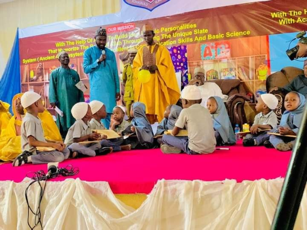 Kinder auf einer Bühne während eines Ereignisses an einer islamischen Schule in Nigeria, mit einem Redner, der ein Mikrofon hält, und einem Banner dahinter, einer Kameraeinstellung rechts und sichtbaren Sofas im Hintergrund.