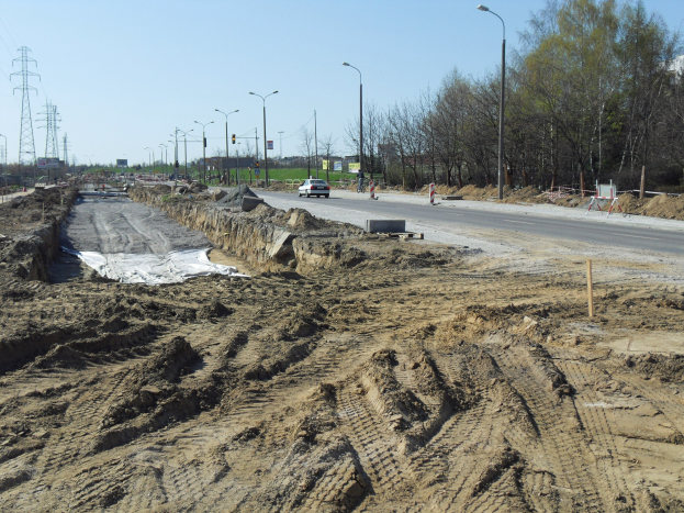 Großer Schutthaufen auf einer Baustelle neben einer Straße mit Fahrzeugen, Laternenmasten, Strommasten mit Kabeln, Bäumen und einem klaren blauen Himmel im Hintergrund.