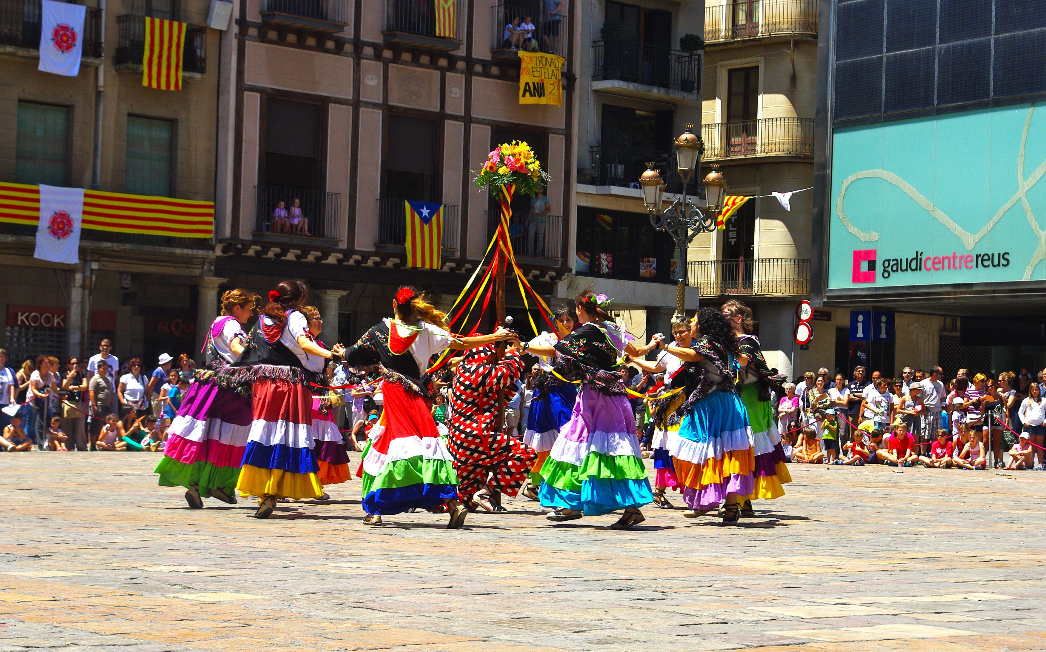 Eine Gruppe von Frauen in bunten Kleidern, die auf der Straße tanzen, umgeben von einer Menge Menschen, mit Gebäuden, Geländern, Balkonen, Fahnen, einem Laternenpfahl und einer Tafel mit Schrift im Hintergrund.