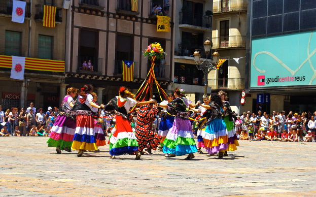 Eine Gruppe von Frauen in bunten Kleidern, die auf der Straße tanzen, umgeben von einer Menge Menschen, mit Gebäuden, Geländern, Balkonen, Fahnen, einem Laternenpfahl und einer Tafel mit Schrift im Hintergrund.