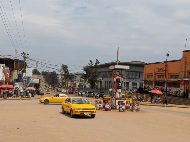 Ein gelbes Taxi fährt auf einer belebten Stadtstraße mit Gebäuden und Passanten mit Regenschirmen auf dem Gehweg und Wolken am Himmel und Stromleitungen oben.