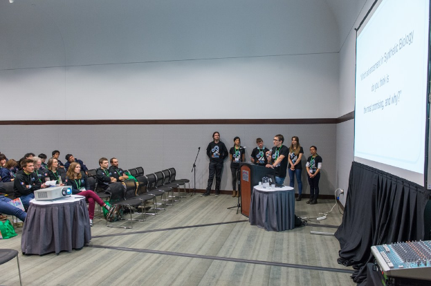 Eine Person, die in ein Mikrofon spricht, bei einem Meeting mit Teammitgliedern, die ID-Karten tragen, einem Pr├Ąsentationsbildschirm und einem Videoplayer in einem leeren Büroraum.