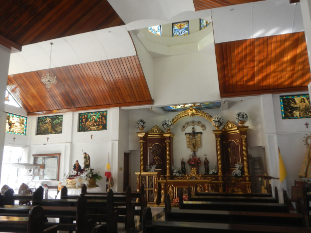 Innenseite einer Kirche mit Holzlehnen, Statuen, Blumenarrangements, Fahnen, gerahmten Fotos an den Wänden und Buntglasfenstern, mit Deckenlampen, die Gemütlichkeit vermitteln.