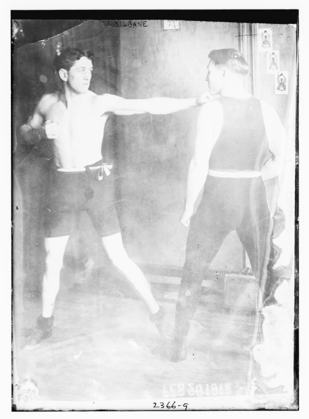 Schwarz-weiß-Foto von zwei Boxern im Ring mit Postern an der Hintergrundwand und Text unten.