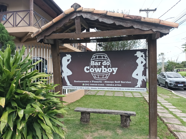 Ein Bar do cowboy Restaurant & Pizzeria mit einer Tafel, die Text und Bilder anzeigt, einer Bank, Gras, Pflanzen, einem Gebäude mit Fenstern, Geländern, Bäumen, Fahrzeugen auf der Straße, Strommasten mit Drähten und einem Himmel im Hintergrund.