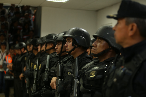 Gruppe von Polizisten in Uniform, die in Formation stehen und Helme sowie Waffen tragen, vor einer Wand und Deckenleuchten im Hintergrund.