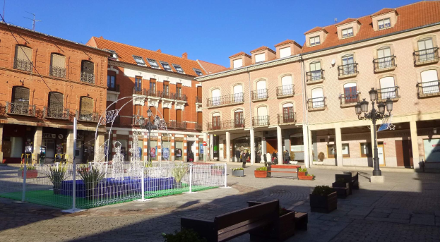 Ein Stadtplatz mit einem zentralen Brunnen, umgeben von Bänken, Topfpflanzen, einem Metallzaun, Straßenlaternen, Menschen, Schildern, Gebäuden mit Fenstern und einem bewölkten Himmel.