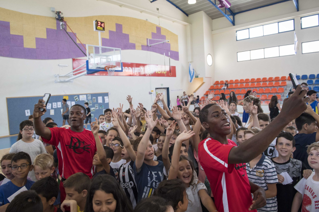 Kinder auf einem Basketballfeld mit Handys stehend, mit einem Korbleger, Basketballnetz, Uhr, an eine Wand geklebten Papieren, Deckenleuchten, Stühlen und Fenstern im Hintergrund.