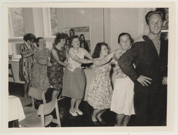 Gruppe von Menschen, die in einem Raum im Stil der 1950er Jahre tanzen, mit Stühlen, einem Tisch mit einer wei├čen Decke, einem Foto Rahmen an der Wand und Fenstern im Hintergrund.