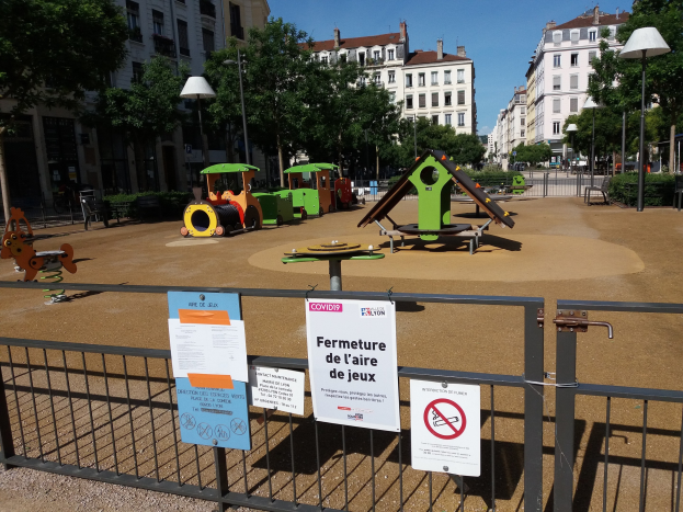 Ein umzäunter Spielplatz in einem städtischen Platz mit Schildern an der Umzäunung, umgeben von Bäumen, Laternenmasten und Gebäuden unter einem sichtbaren Himmel.