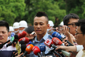 Ein Polizeibeamter von Reportern mit Mikrofonen und Aufzeichnungsgeräten umgeben, mit Bäumen im Hintergrund.