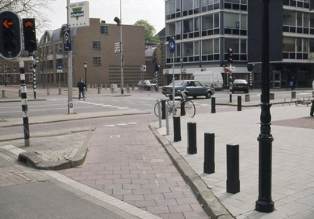 Eine Stadtstraße mit einem geparkten Fahrrad, fahrenden Fahrzeugen, Fußgängern auf dem Gehweg, Strommasten, Verkehrsampeln, Straßenschildern, Bäumen, Gebäuden mit Fenstern und einem klaren blauen Himmel.