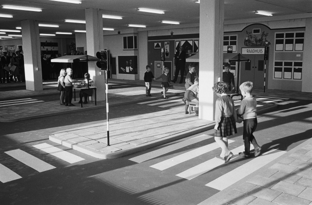 Schwarz-weiß-Foto von Kindern, die eine Straße an einer Ampelübergang mit nahestehenden Passanten überqueren, mit einer Verkehrsampel im Vordergrund und Hintergrundsäulen mit Deckenleuchten.