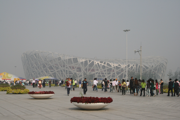 Olympisches Stadion in Peking, China mit Menschen, die herumlaufen, Blumenpflanzen im Vordergrund, Laternenpfähle, Bäume und einen klaren blauen Himmel im Hintergrund.