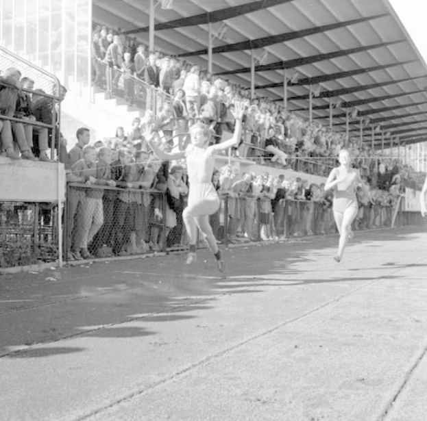 Zwei Frauen bei einem Weitsprung auf einer Laufbahn, eine schwebt in der Luft, mit Zuschauern im Hintergrund und einem Zaun und einer Hütte sichtbar.