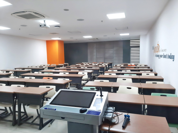 Ein Klassenzimmer mit Tischen und Stühlen in Reihen, einem deckengehängten Projektor und Gegenständen auf den Tischen, mit Text an der rechten Wand, der auf eine Privatschule in Singapur hinweist.