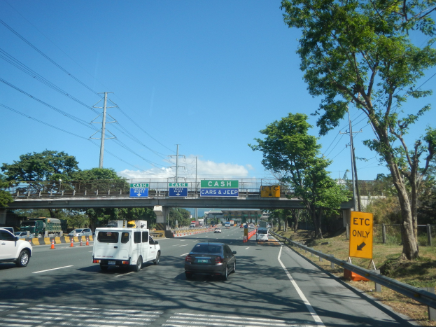 Eine Autobahn mit fahrenden Autos, eine Brücke im Hintergrund, Strommasten mit Drähten, Verkehrsschilder, Wärner, Gras, Bäume und ein bewölkter Himmel.