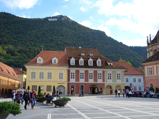 Menschen gehen in einem Stadtplatz umgeben von Gebäuden mit Fenstern, Laternenmasten und Topfpflanzen, mit einem Hügel und bewölktem Himmel im Hintergrund und Text auf einem Gebäude.