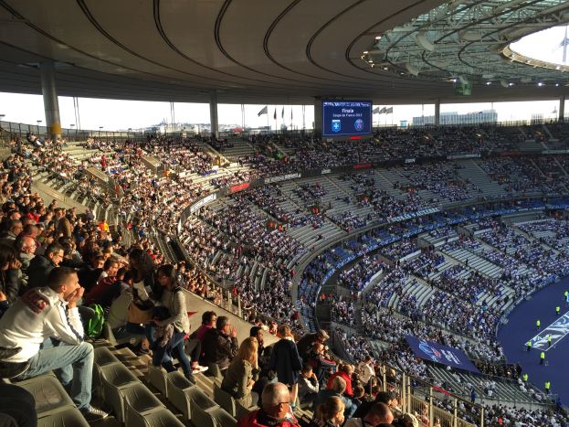 Große Menschenmenge in einem Stadion bei einem Fußballspiel, mit einer Bühne rechts, Fahnen, Stangen, einem Bildschirm und dem Allianz Stadion in München im Hintergrund.