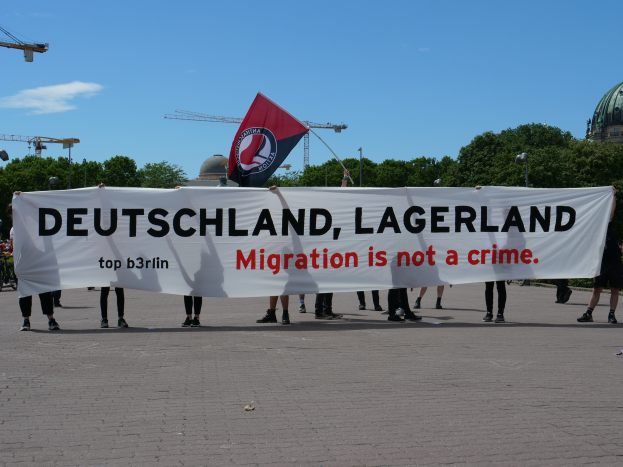 Eine Gruppe von Menschen hält ein Transparent mit der Aufschrift "Deutschland, Lagerland Migration ist kein Verbrechen" und eine Fahne, sie stehen auf dem Boden mit Straßenlaternen, Straßenlaternen, Bäumen, Kränen, einem Gebäude und einem bewölkten Himmel im Hintergrund.