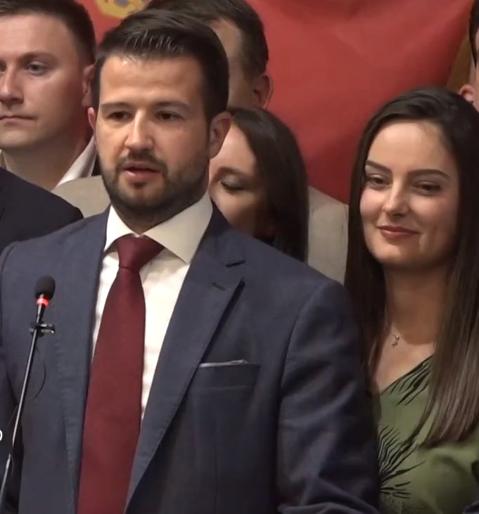 Ein Mann im Anzug spricht in ein Mikrofon an einem Podium mit einem Banner dahinter und spricht zu einer Menge.