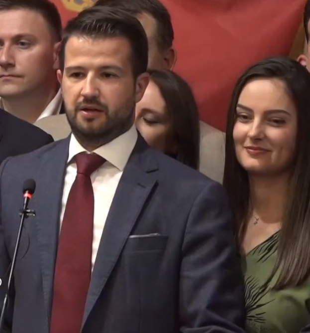 Ein Mann im Anzug spricht in ein Mikrofon an einem Podium mit einem Banner dahinter und spricht zu einer Menge.