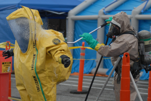 Zwei Personen in Schutzanzügen sprühen sich gegenseitig mit Wasser voll, eine hält dabei eine Sprühpistole, während die andere im Vordergrund steht, mit blauen Zelten und einer Straße im Hintergrund.