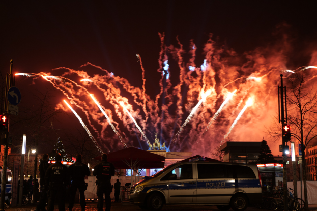 Polizeiauto vor Menschenmenge bei Neujahrsfeierlichkeiten in Berlin geparkt, mit farbigen Feuerwerken am Himmel.