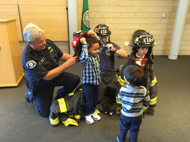 Eine Gruppe von Kindern in Feuerwehruniformen und ein Mann in Feuerwehruniform versammeln sich um einen Helm, mit einem Feuerwachen-Interieur im Hintergrund.