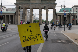 Eine Gruppe von Radfahrern mit Helmen fährt vor dem Brandenburger Tor in Berlin, Deutschland, vorbei, während einer ein gelbes Schild hält, unter einem klaren blauen Himmel mit Gebäuden und Bäumen im Hintergrund.