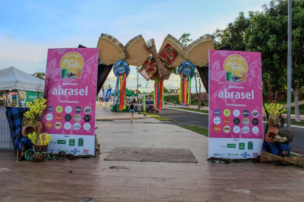 Gruppe von Menschen vor einem "Abrasel"-Schild auf einem Festival mit Bannern, einem sitzenden Menschen, einem Besenstiel, Topfpflanzen, einem Tisch, einem Zelt, Pfosten, Gras, Bäumen, dekorativen Gegenständen, einer Straßenlaterne, Fahrzeugen, Gebäuden und einem bewölkten Himmel.