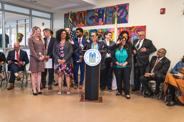 Gruppe von Menschen in einem Raum mit einem Podium, einem Mikrofon und einer Tafel mit der Aufschrift "Sancho-Stadtrat tritt dem Stadtrat bei", begleitet von Flaggen, einem Feuerlöscher und Deckenleuchten.