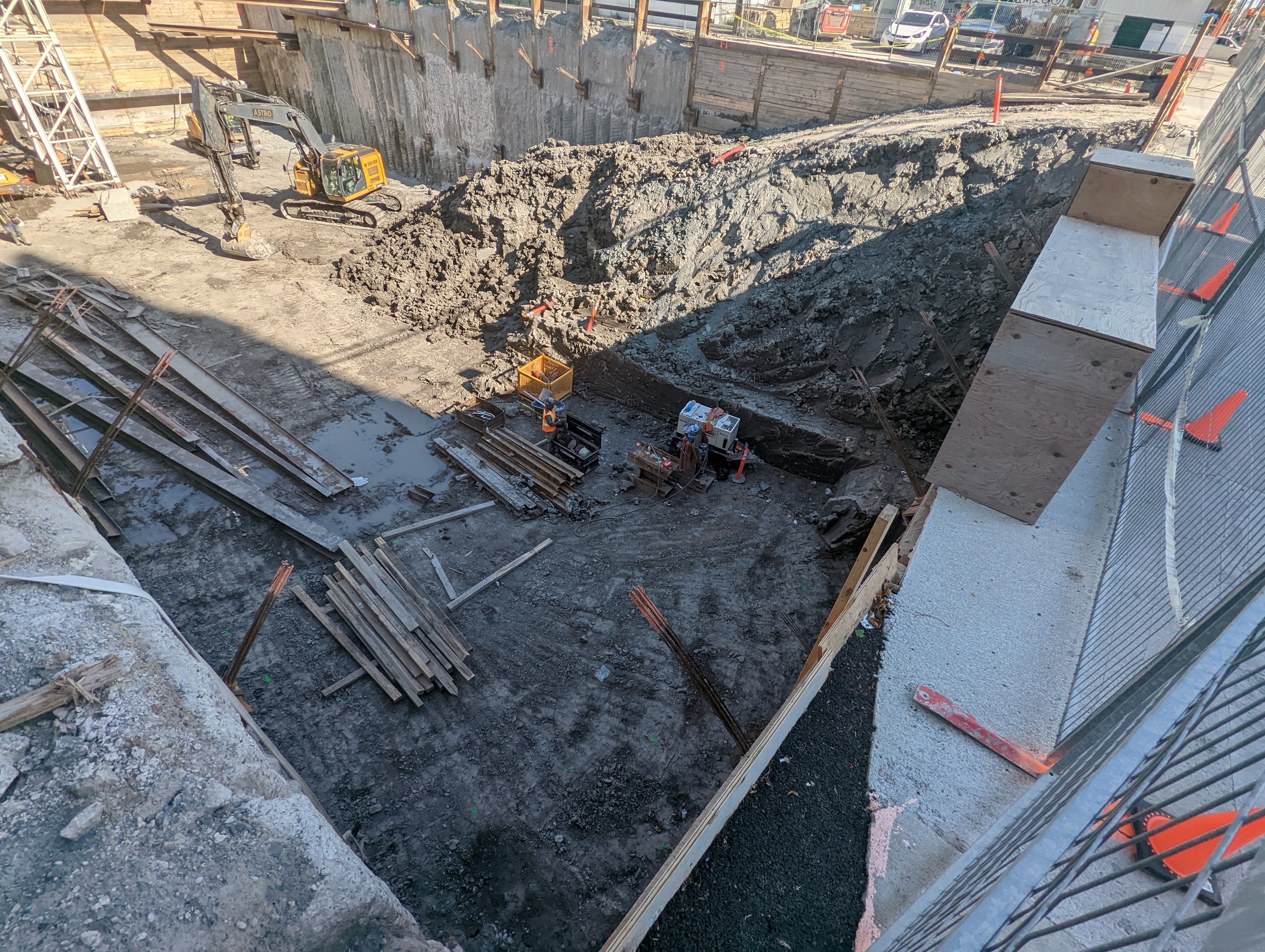 Baustelle mit einem Erdwall, Kränen, Holzpfählen, einem Maschendrahtzaun auf der rechten Seite und Hintergrundelementen wie Gebäuden, Fahrzeugen, Pfählen, Bäumen und Himmel.